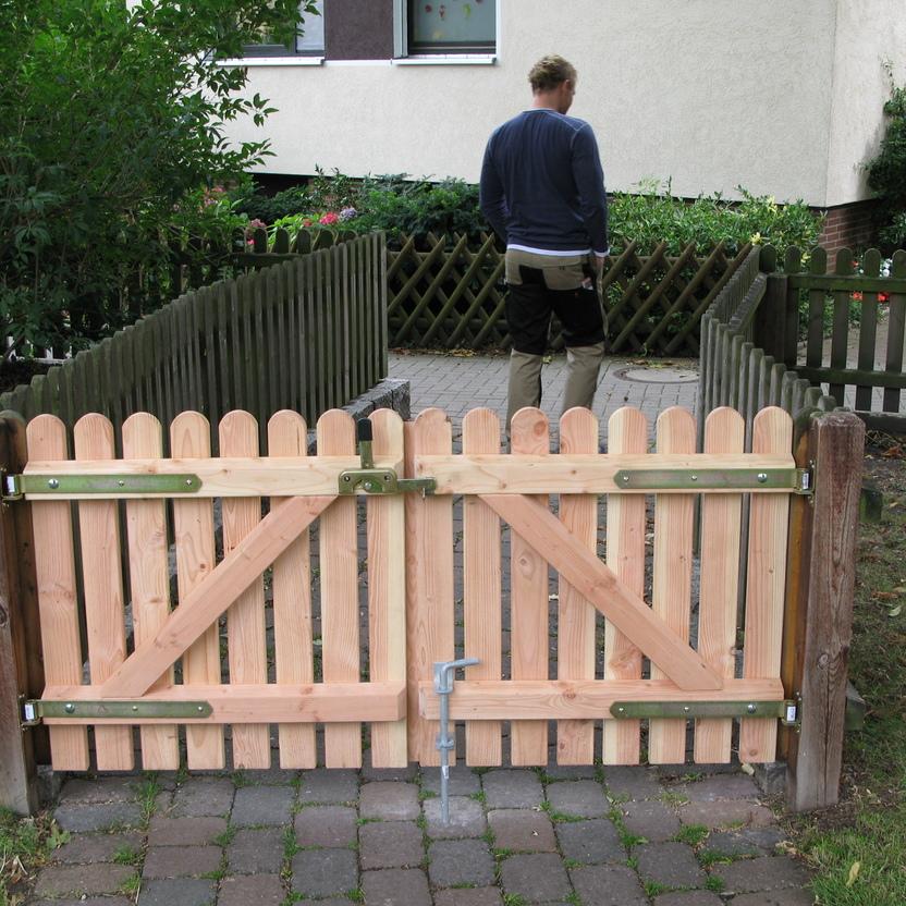 Robustes Holzgartentor Ein stabiles, doppeltes Holzgartentor mit Metallbeschlägen an einem gepflasterten Zugang, ein Mann im Hintergrund geht den Weg entlang.