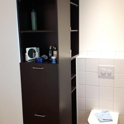 Maßgefertigtes Badmöbel neben einer Toilette Modernes Badezimmer mit einem hohen, dunklen Badmöbel nach Maß, das an der Seite offene und geschlossene Aufbewahrungsbereiche bietet, neben einer Wandtoilette.