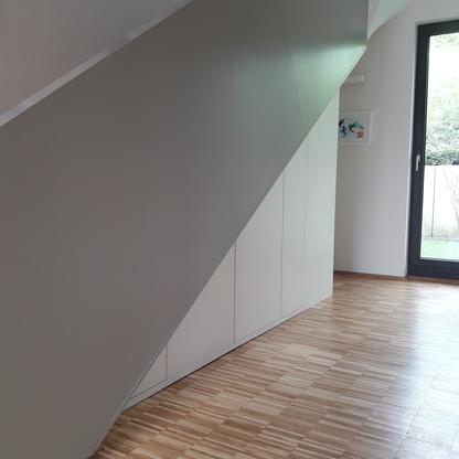 Maßgeschneiderter Einbauschrank unter einer Schräge Ein langer, weißer Einbauschrank passt nahtlos unter die Schräge einer modernen Treppe mit Holzparkettboden und hellem Tageslicht.