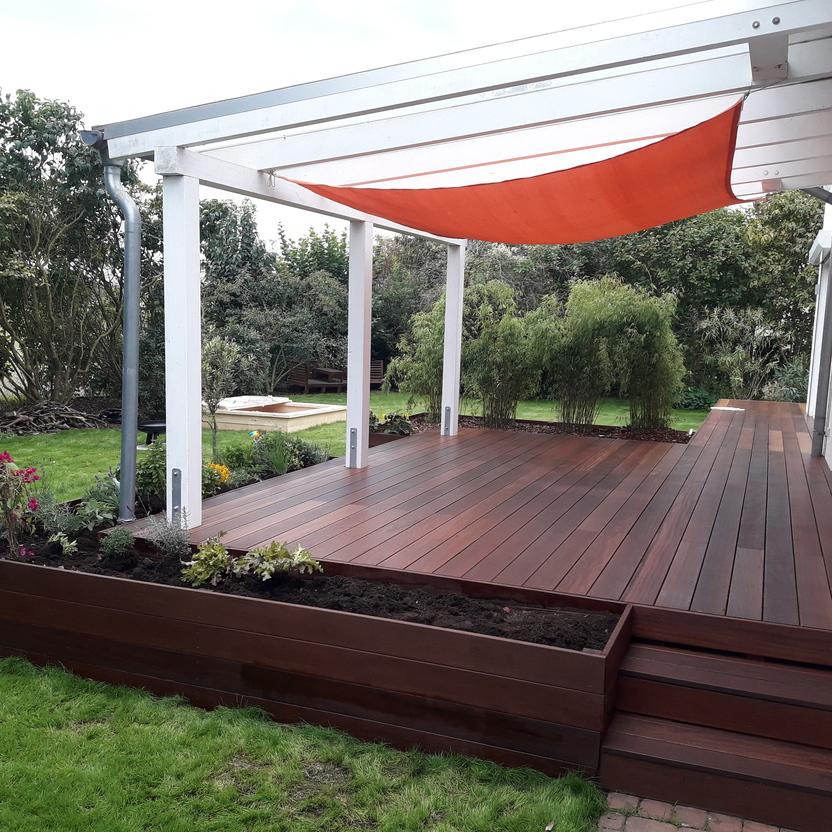 Maßgefertigte Holzterrasse mit Überdachung Elegante Terrasse nach Maß mit dunklem Holzboden und weißer Überdachung, ausgestattet mit einem orangefarbenen Sonnensegel, umgeben von einem gepflegten Garten.