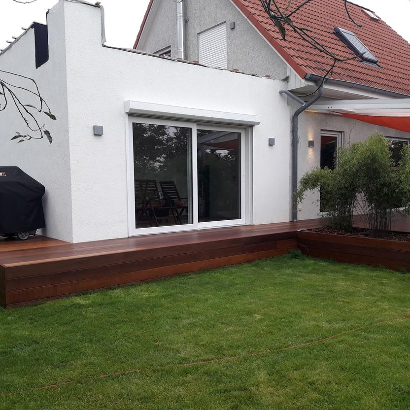 Maßgefertigte Terrasse an einem modernen Haus Eine Terrasse nach Maß aus dunklem Holz, angrenzend an ein weißes Haus mit großen Fenstern und einem orangefarbenen Sonnensegel. Ideal für den Außenbereich.