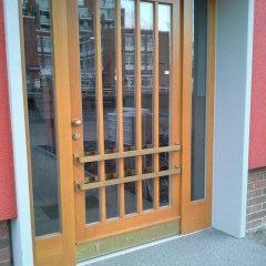 Holztür nach Maß mit Gitterdesign Maßgefertigte Holztür mit Sicherheitsgitter und Glaseinsätzen, umgeben von einem grauen und roten Mauerwerk, bietet einen geschützten Eingang.