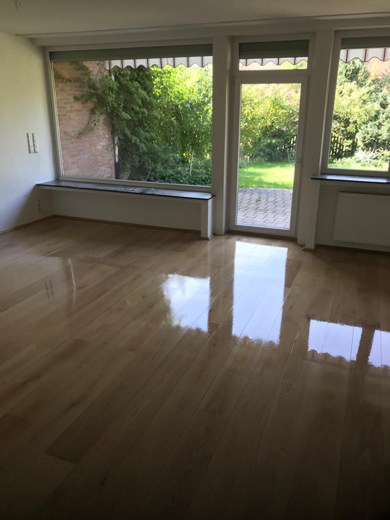 Helles Wohnzimmer mit großem Fenster und Gartenzugang Geräumiges, leeres Wohnzimmer mit glänzendem Holzboden, großen Fenstern und einer Glastür, die einen Blick auf den grünen Garten freigibt.